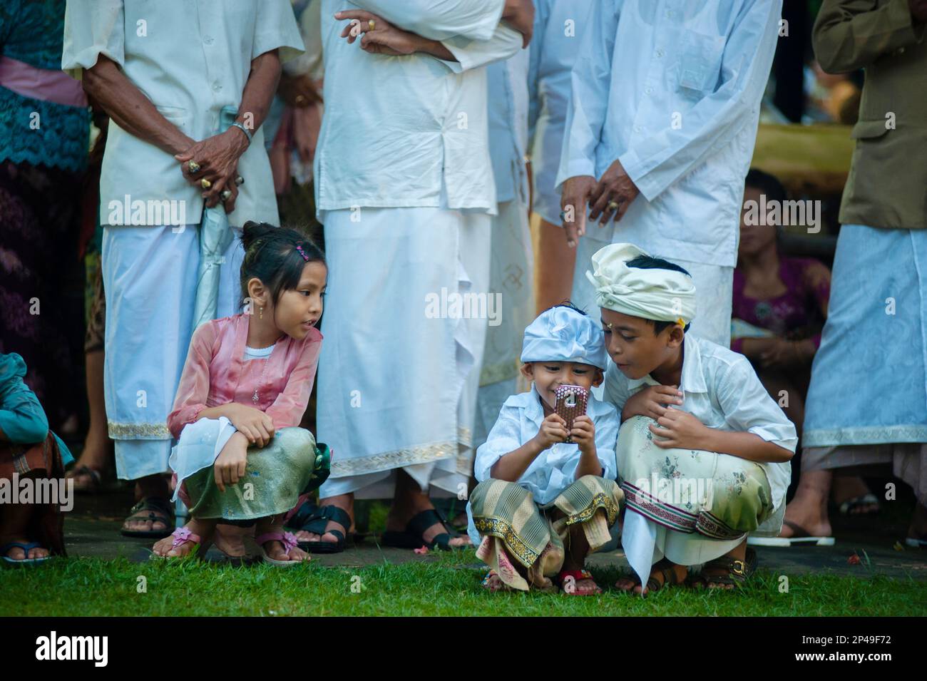Ragazza che guarda due ragazzi che giocano con il telefono cellulare con adulti in piedi dietro durante il festival Siat Sampian (guerra delle foglie di cocco), pura Samuan Tiga, Ubud, Foto Stock