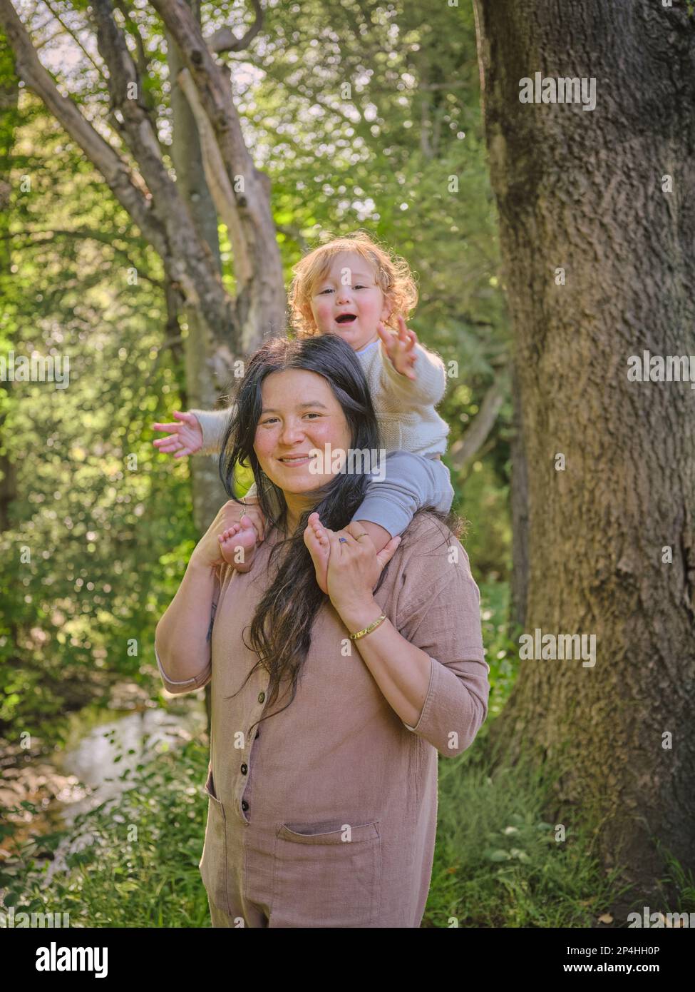 Figlia si siede sulle spalle delle madri vicino al torrente in un ambiente boscoso Foto Stock