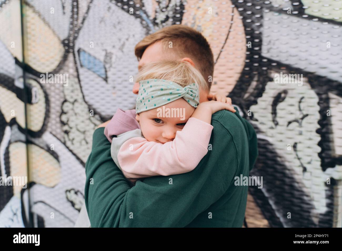 Papà in felpa con cappuccio che abbraccia una figlia di due anni di fronte ai graffiti Foto Stock
