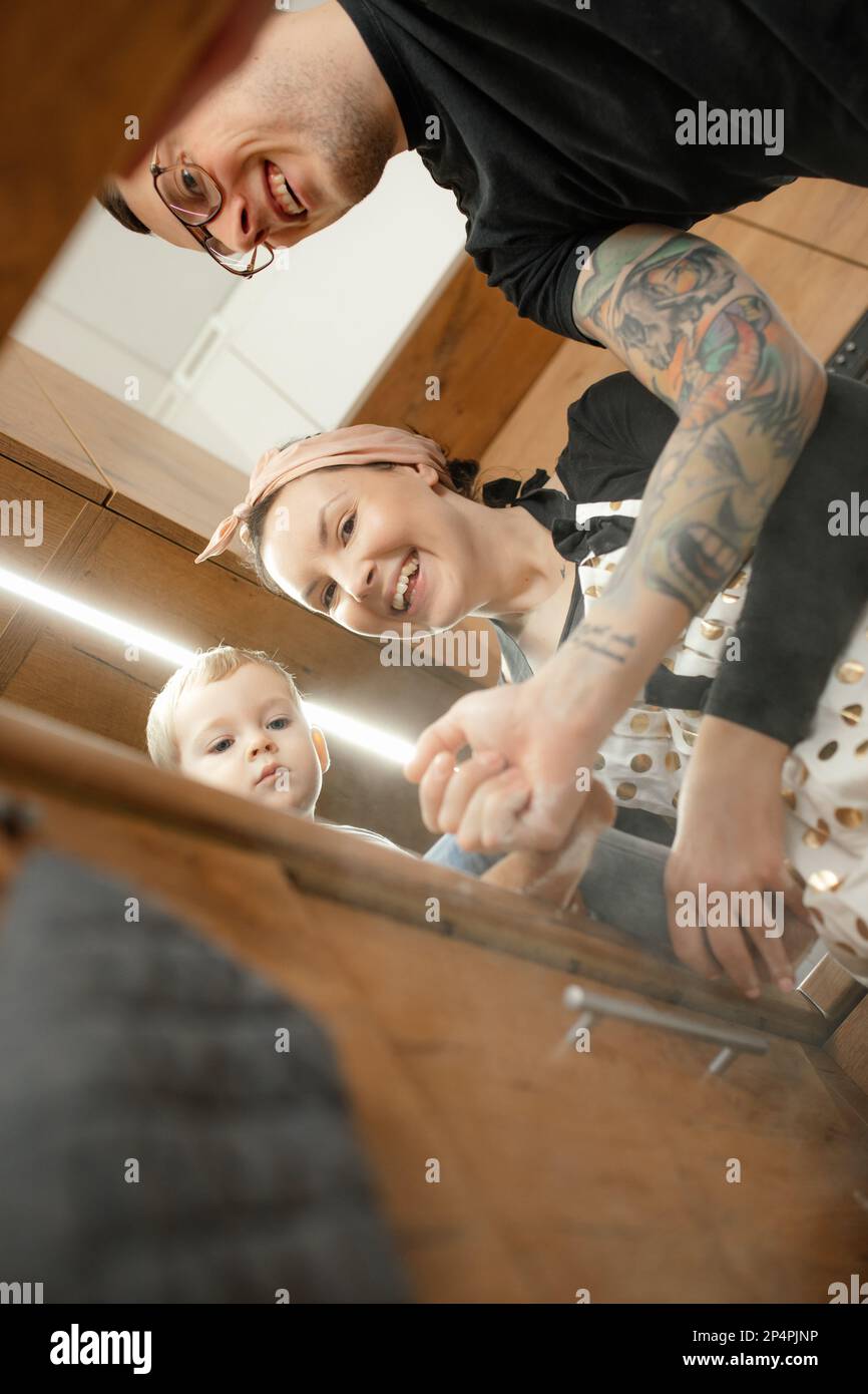 Foto verticale di donna malinetta ridente, uomo con manica di tatuaggio e ragazzino che guarda giù alla macchina fotografica in cucina Foto Stock
