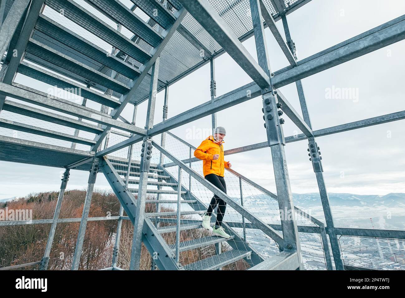 Corridore sorridente vestito con abiti sportivi color arancione chiaro softshell che scendono da enormi scale industriali in acciaio nel freddo giorno d'inverno. Persone sane l Foto Stock