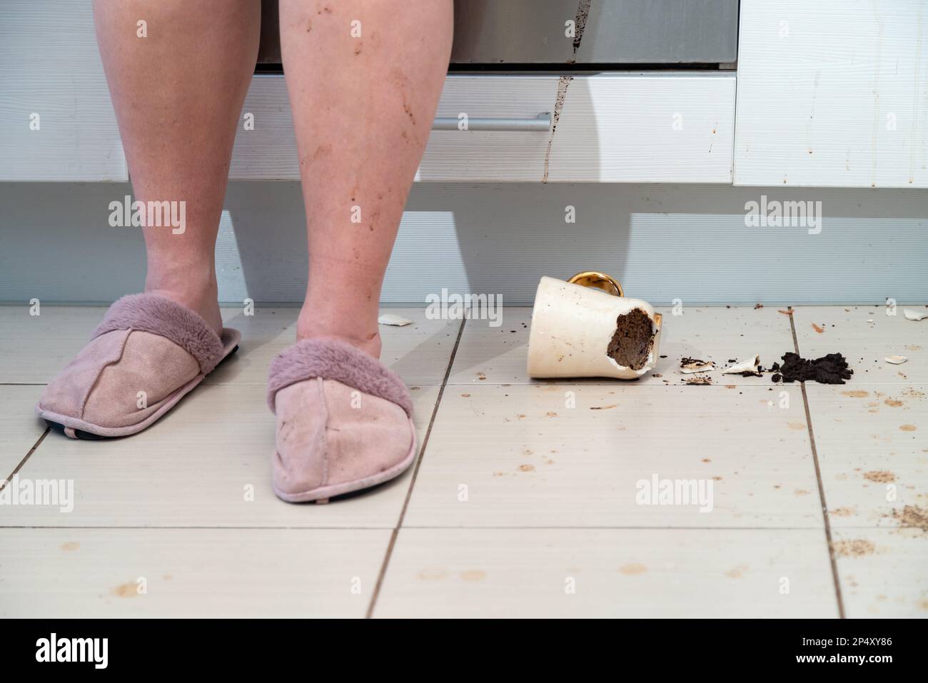Donna in piedi accanto alla tazza da tè rotta che si trova sul pavimento della cucina, tazza da caffè e caffè macinato in tutto il mattonelle. Foto Stock