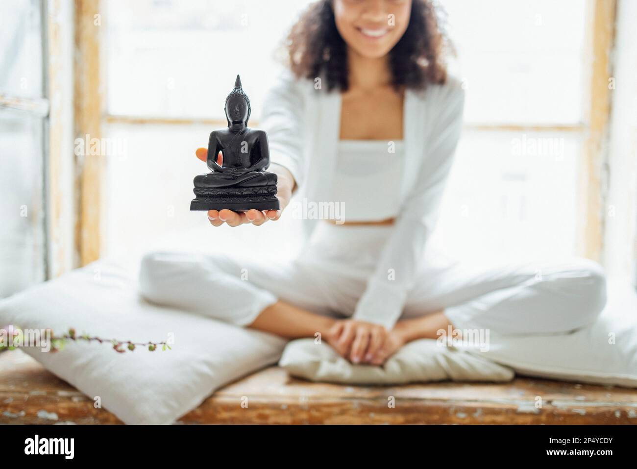 Primo piano di una statua di Buddha sulle mani di una ragazza dalla pelle scura. Giovane donna afroamericana sorridente in abiti bianchi seduta in posizione lotus su vittoria Foto Stock