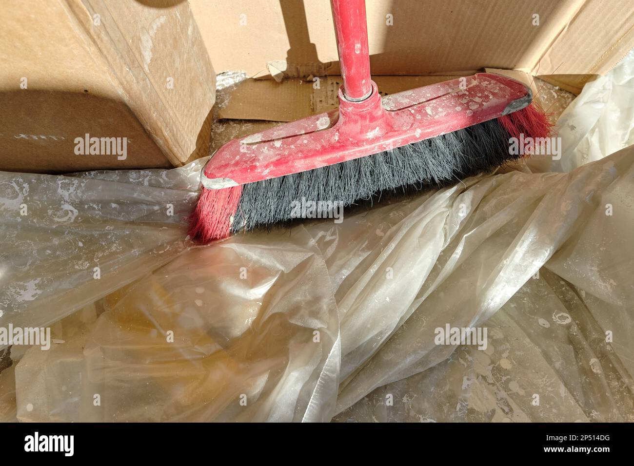Pavimento in legno ricoperto di lamina trasparente con macchie e sbavature di vernice bianca in preparazione alla verniciatura delle pareti. Scopa rossa accanto al cartone Foto Stock
