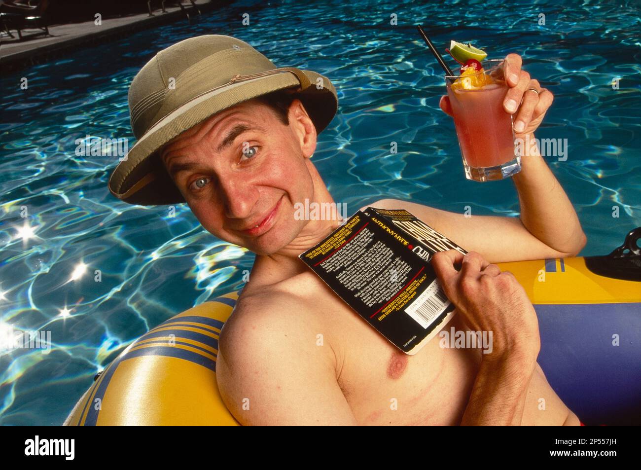 Uomo che indossa un cappello da safari con un libro cartaceo e un drink mentre galleggia in piscina in una zattera Foto Stock
