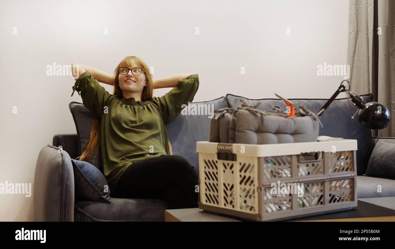 donna con i capelli di zenzero e gli occhiali poggiati sul divano e godendosi la sua nuova casa. Foto di alta qualità Foto Stock