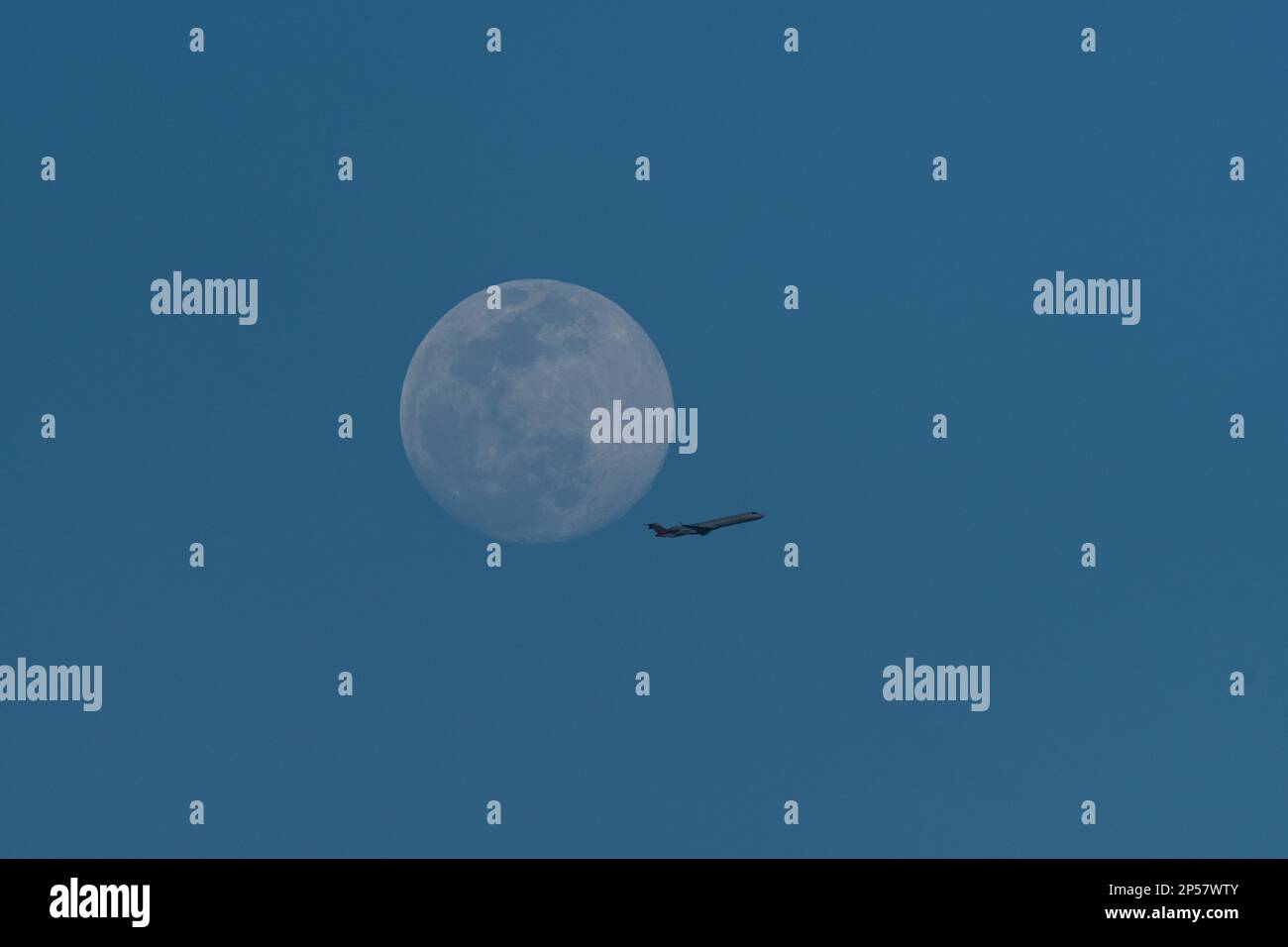 Un jet passeggeri che passa dalla luna quasi piena in alto in un cielo limpido e blu diurno. Foto Stock