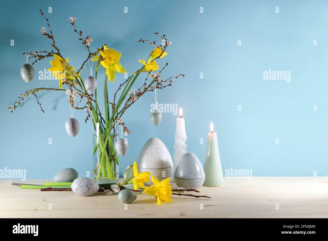 Decorazione stagionale delle vacanze con candele, un bouquet di narcisi e rami di primavera in un vaso di vetro e uova di Pasqua grigie artificiali su un legno chiaro Foto Stock