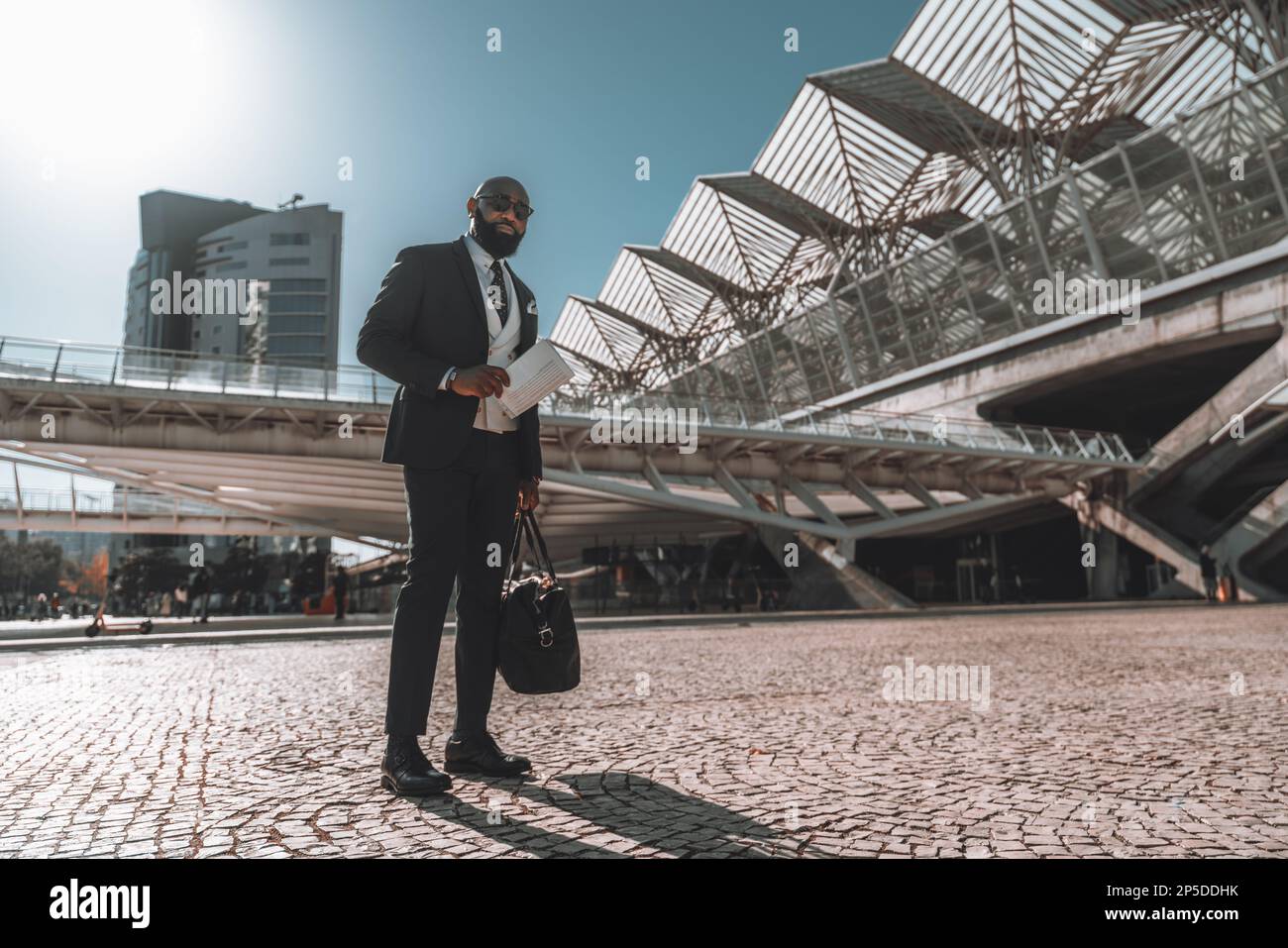 Un uomo nero barbuto su un marciapiede portoghese vicino alla stazione ferroviaria di Oriente, tenendo il quotidiano e una borsa per il fine settimana. Sta indossando un vestito completo A. Foto Stock