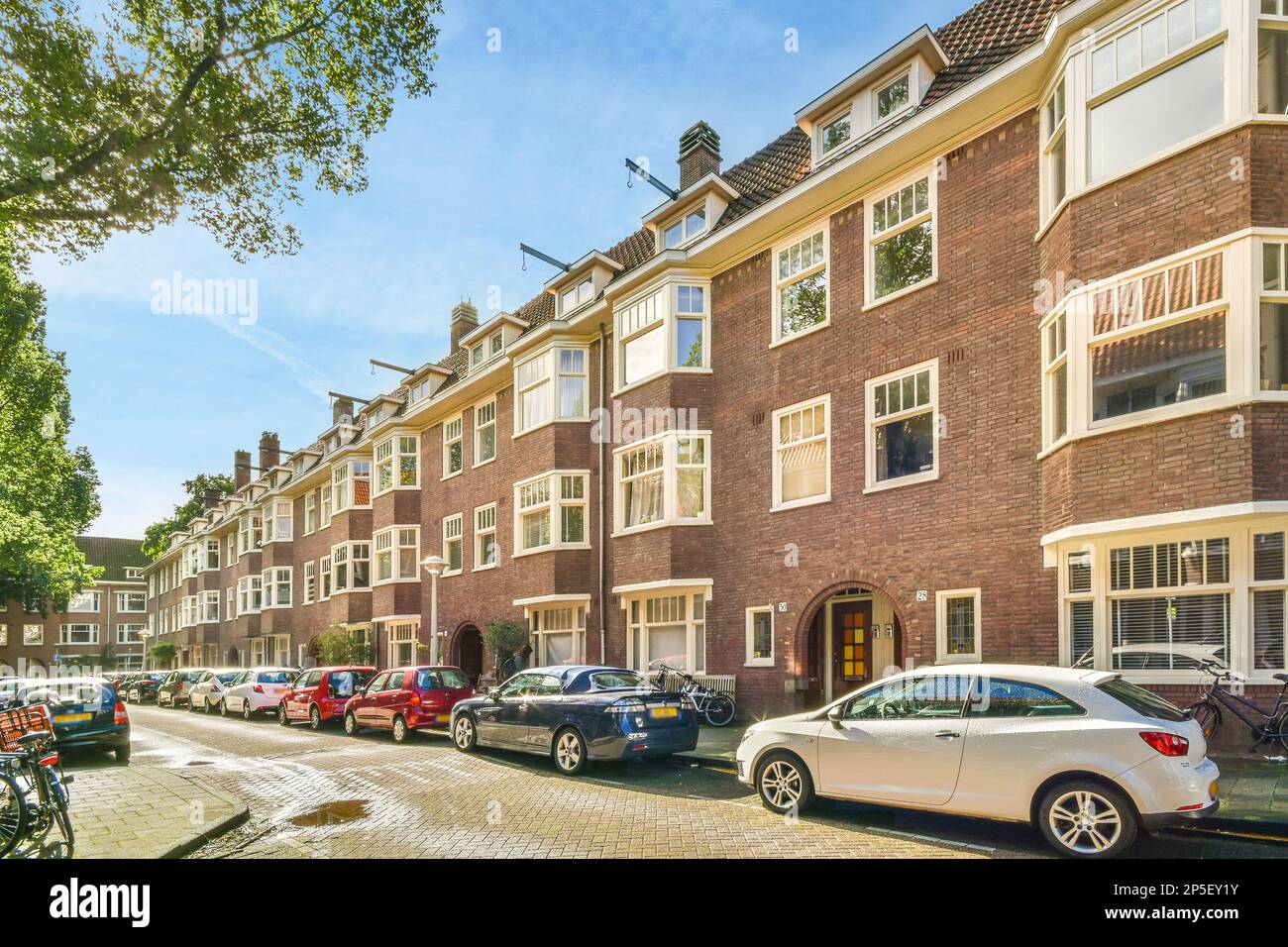 Amsterdam, Paesi Bassi - 10 aprile, 2021: Auto parcheggiate sul lato di una strada di fronte a una fila di edifici in mattoni con finiture bianche e finestre Foto Stock