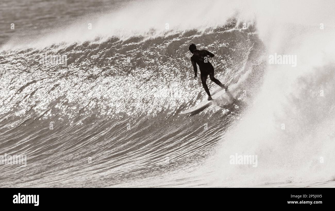 Surf surfista irriconoscibile in cappuccio e muta posteriore azione seppia tono fotografia cavalca fredda offshore ventoso oceano onda Foto Stock
