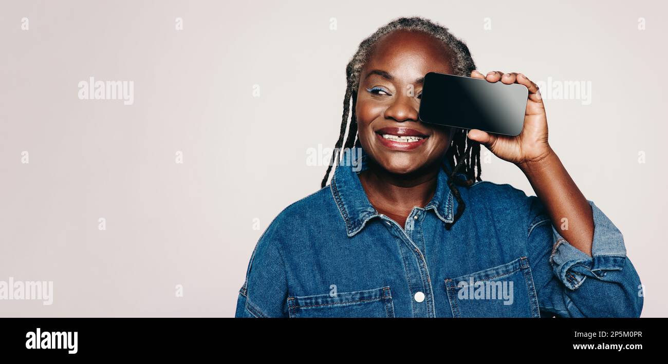 Donna allegra che guarda via con un sorriso mentre tiene uno smartphone davanti al suo occhio. Donna felice matura con i Dreadlocks che portano una giacca di denim e. Foto Stock