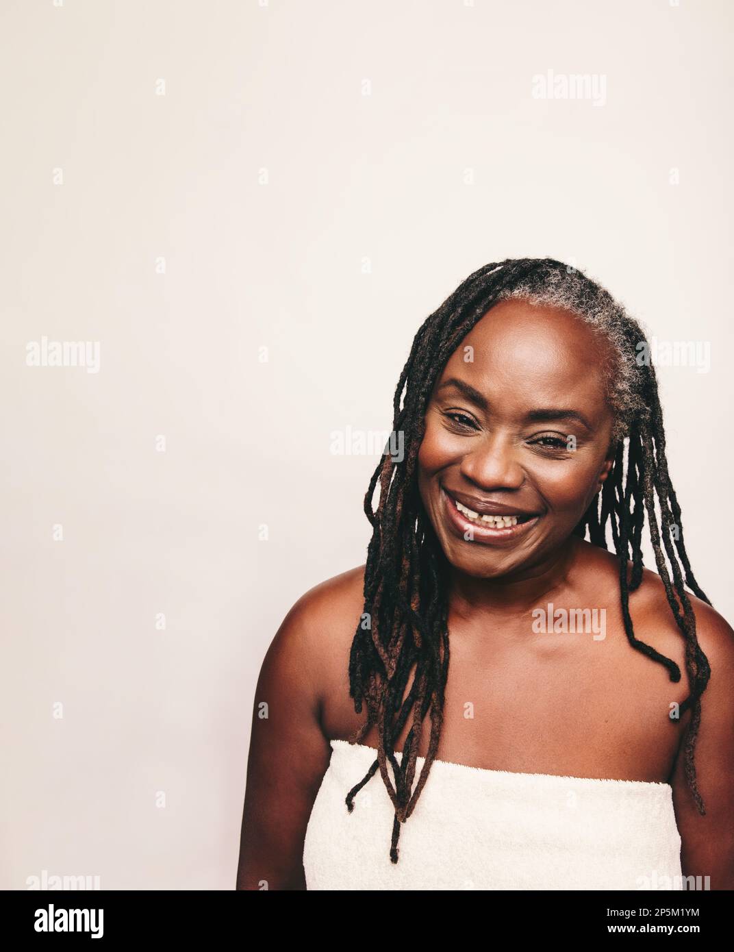 Donna nera matura sorridente alla macchina fotografica mentre avvolta in un asciugamano da bagno. Donna rinfrescata con greggi in piedi contro uno sfondo studio. Buon mi Foto Stock