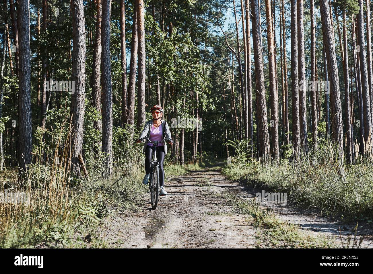 Donna attiva in bicicletta su strada forestale. Donna in bicicletta in fuoristrada percorso durante il giorno delle vacanze estive. Donna che indossa casco e guanti da bicicletta Foto Stock