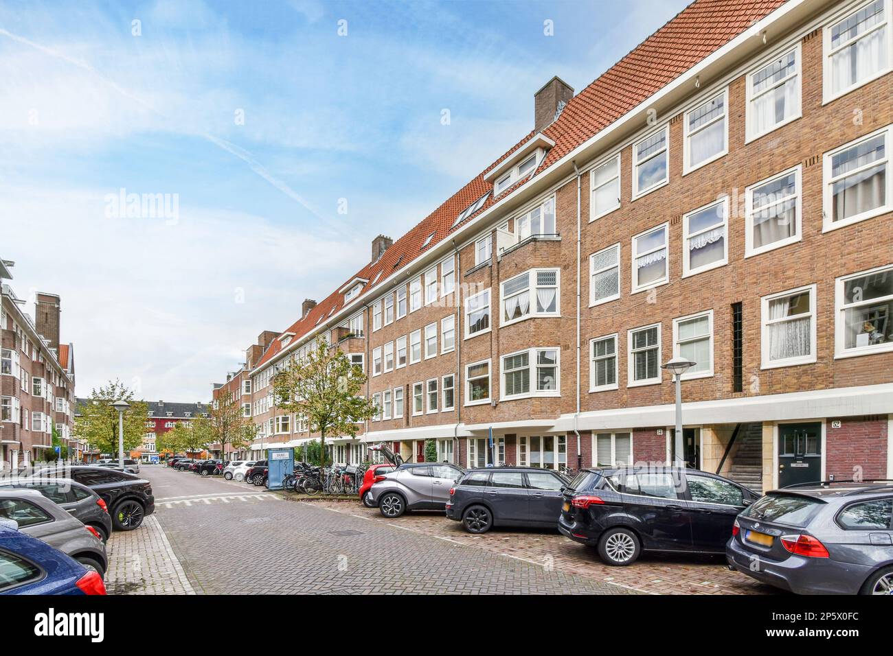 Amsterdam, Paesi Bassi - 10 aprile 2021: Alcune auto parcheggiate sul lato di una strada di fronte ad un edificio in mattoni con finestre bianche e tetto rosso Foto Stock