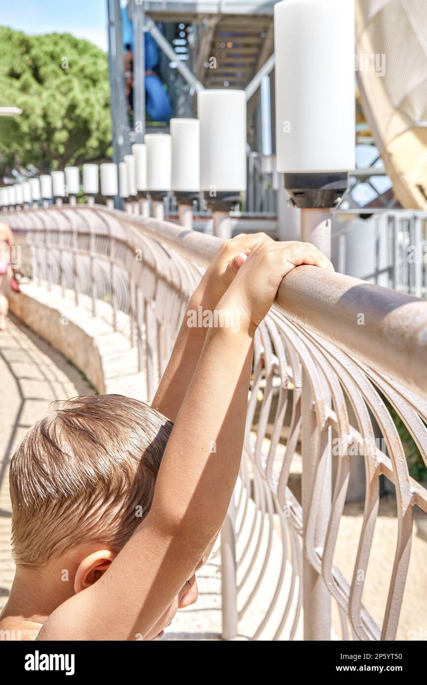 Il ragazzo biondo con i capelli bagnati tiene la ringhiera vicino al grande scivolo nel parco acquatico nelle giornate di sole. Il preschooler ama le vacanze estive al primo piano del resort Foto Stock