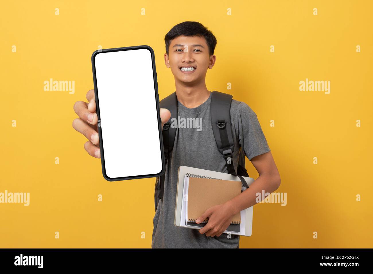 Sorridente giovane studente universitario con libri e zaino isolato su sfondo giallo mostrando grande smartphone con schermo bianco in mano. Pubblicità Foto Stock