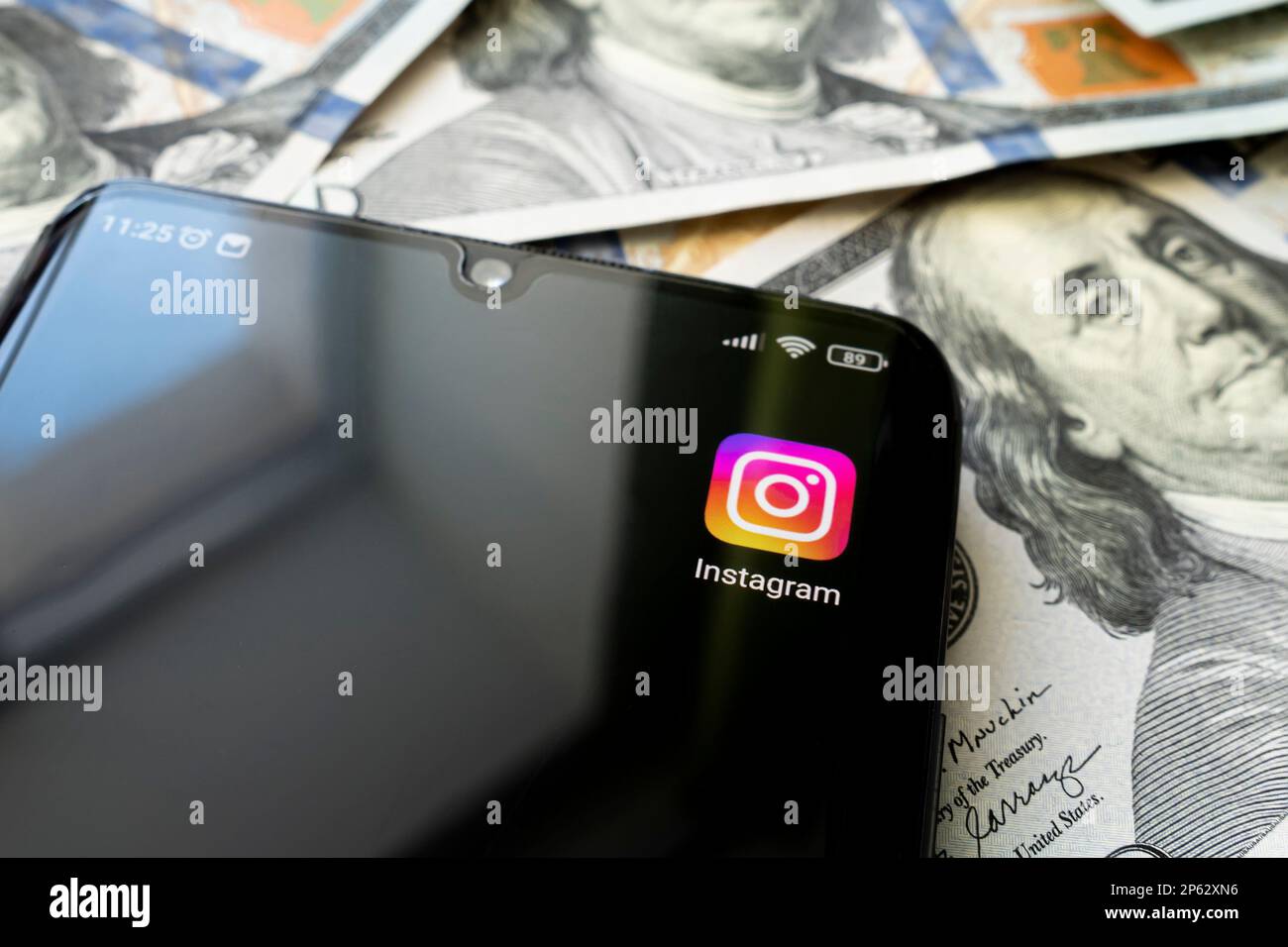 Schermo dello smartphone con l'app Instagram e molte banconote da cento dollari. Concetto di business e social networking. l'icona dell'applicazione sulla schermata nera Foto Stock