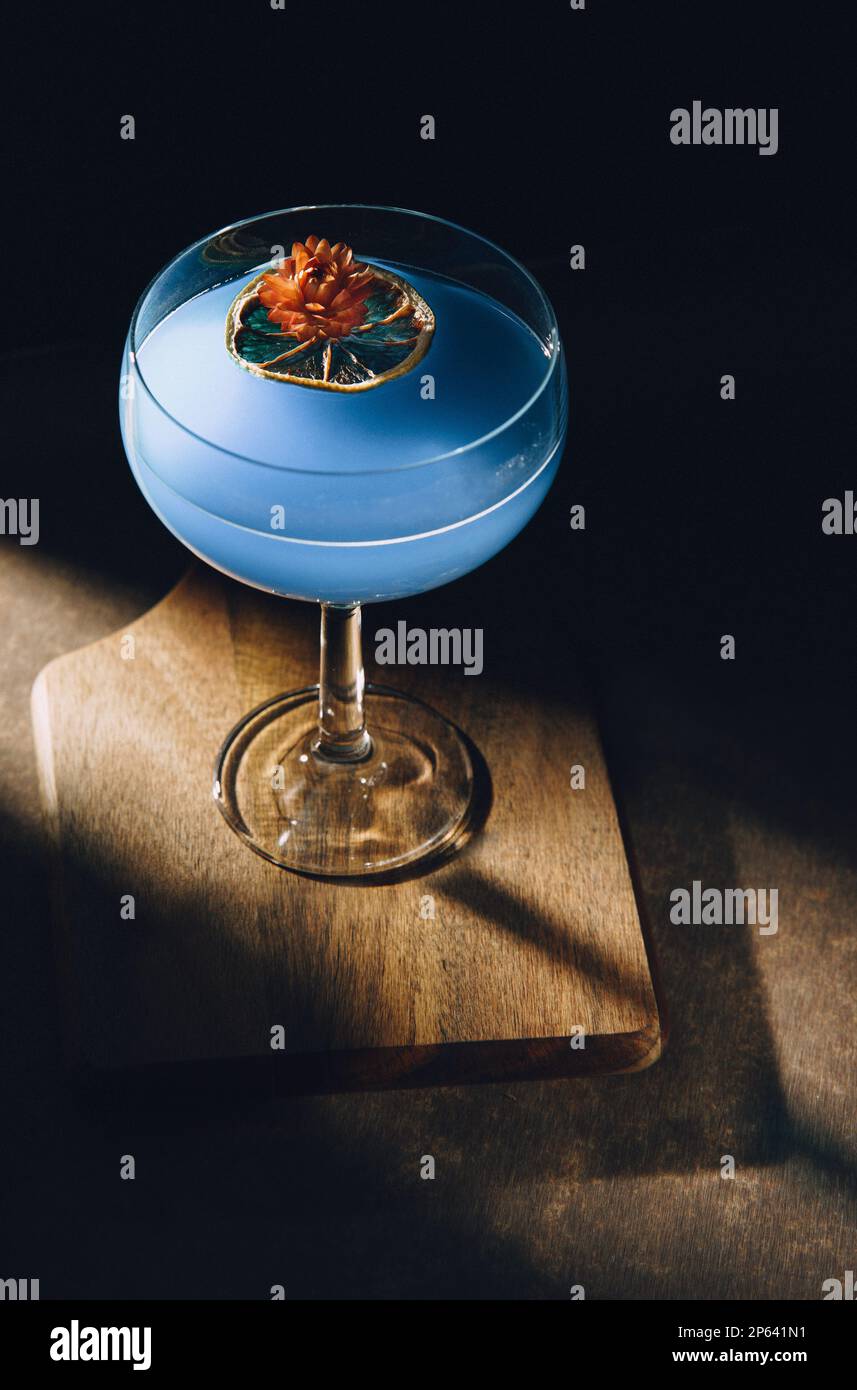 un bel bicchiere arrotondato con liquido blu, una fetta secca di limone, un fiore secco e un noir di umore scuro con un'ombra forte. Foto Stock un bel bicchiere arrotondato con liquido blu, una fetta secca di limone, un fiore secco e un noir di umore scuro con un'ombra forte. Foto Stock