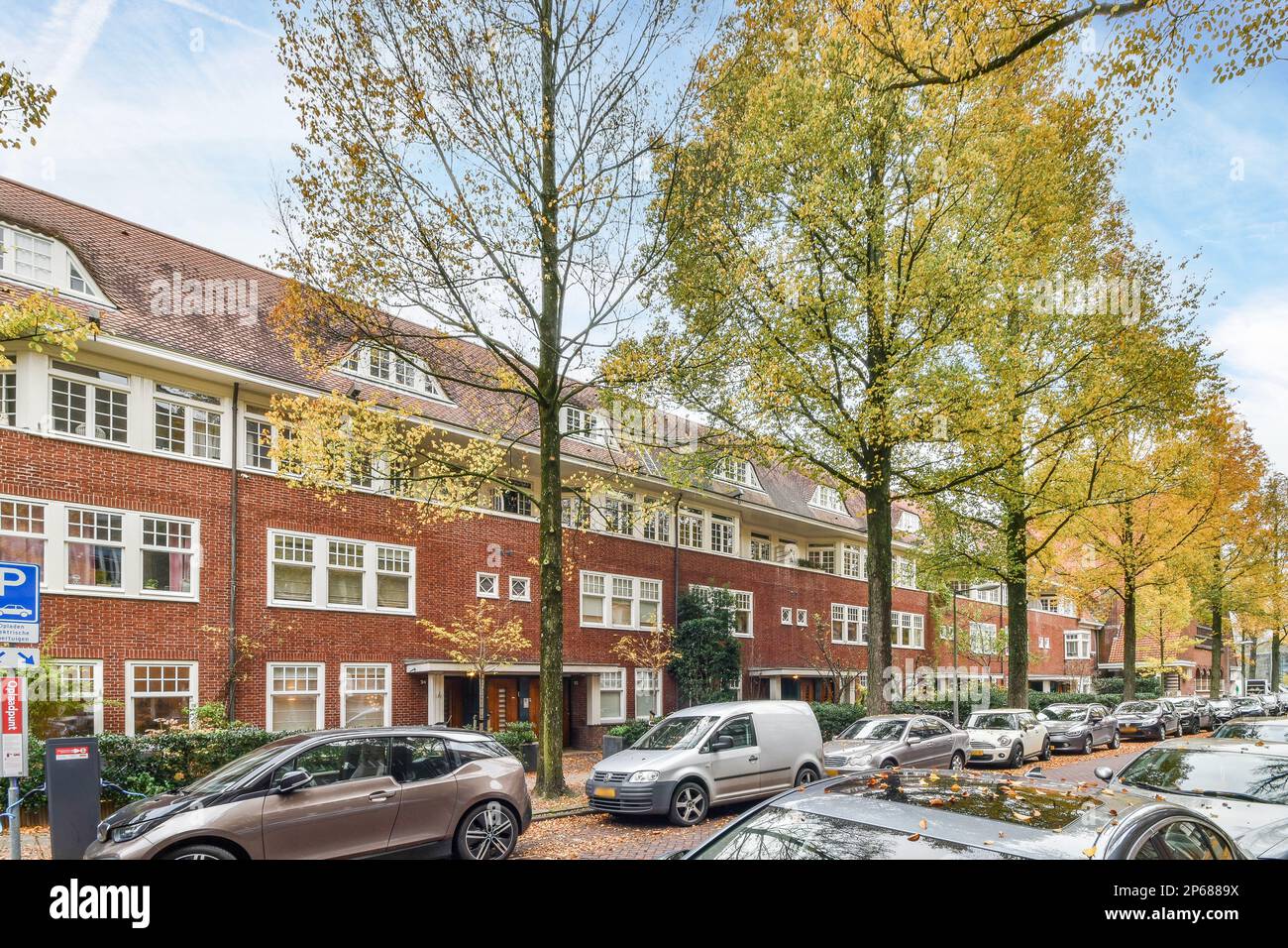 Amsterdam, Paesi Bassi - 10 aprile, 2021: Auto parcheggiate sulla strada di fronte ad un edificio in mattoni rossi con finestre bianche e alberi che fiancheggiano la strada Foto Stock