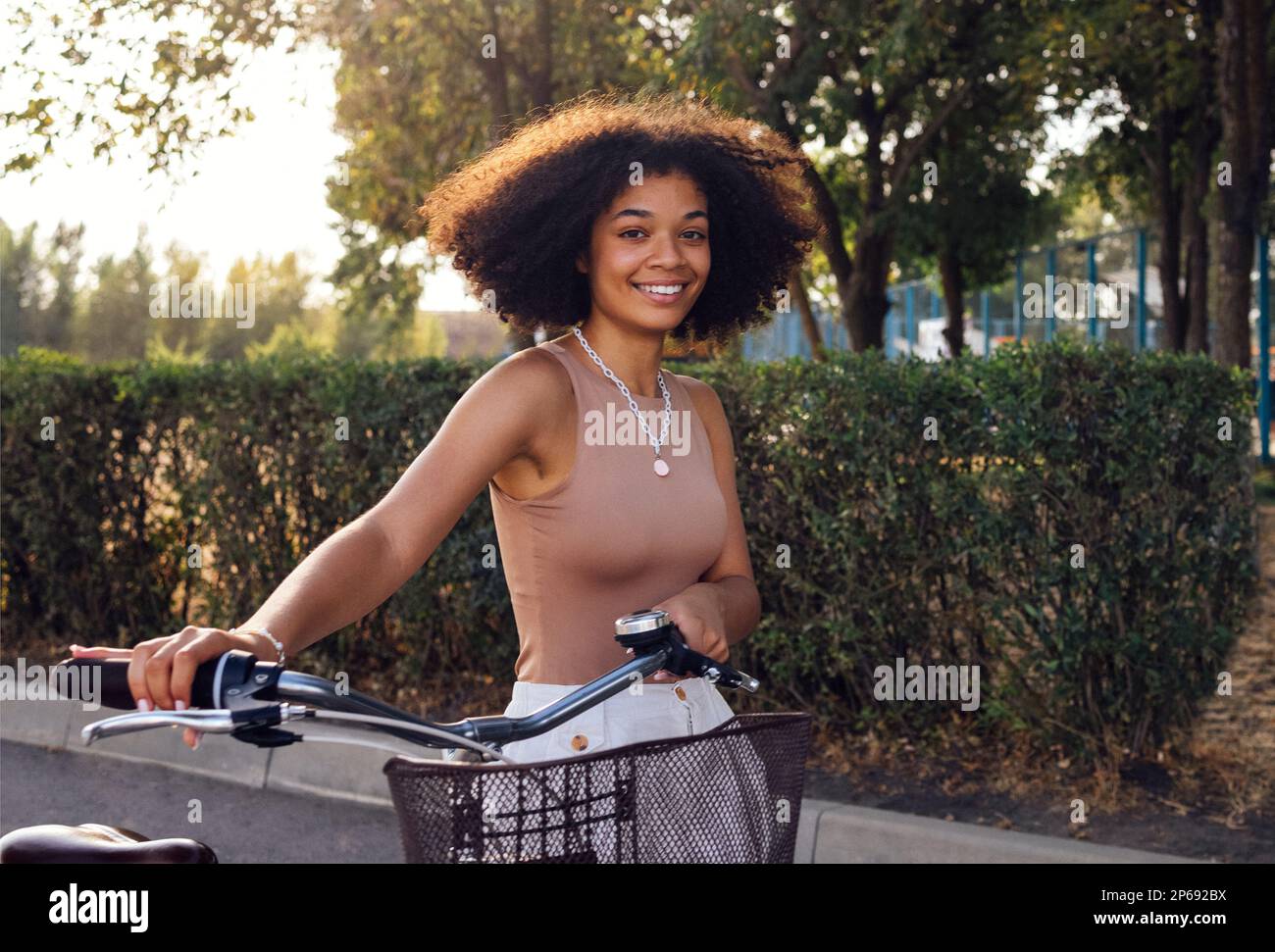 Giovane donna con pelle scura sta guidando la sua bicicletta. Giovane e positiva afro ragazza americana in tessuto casual sorride dolcemente e ha un divertimento. Strada cittadina AN Foto Stock