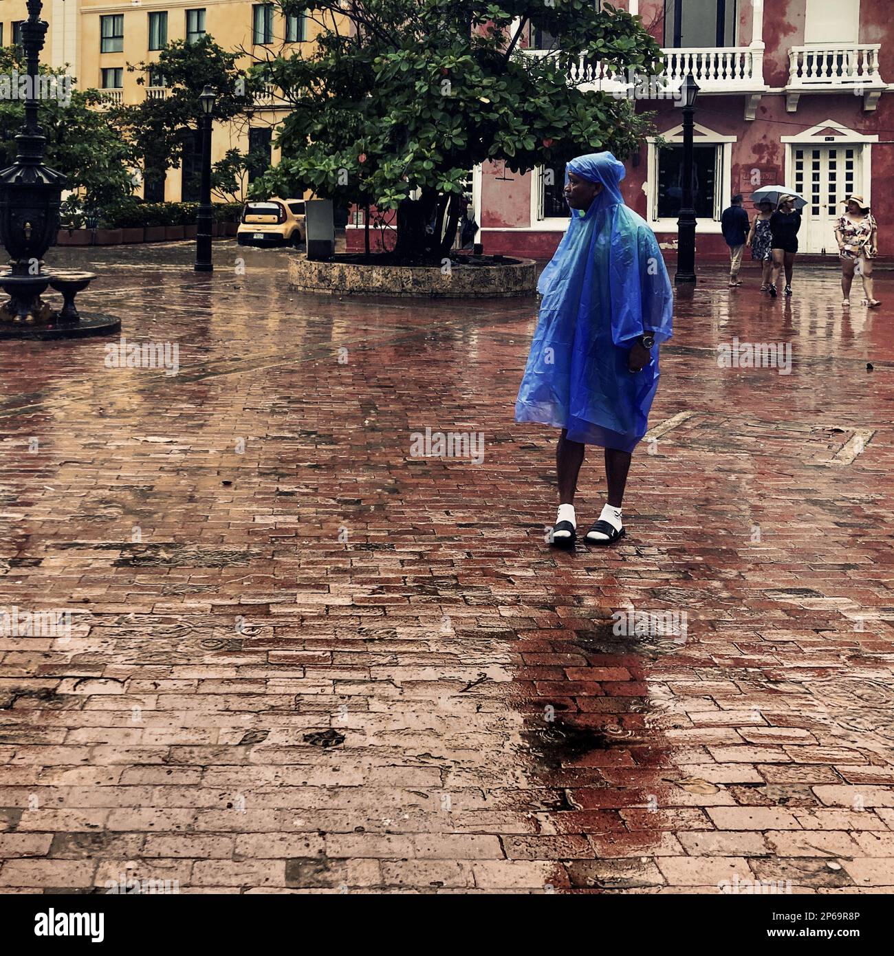 Un afro-colombiano, indossando un impermeabile, cammina in una piazza acciottolata sotto la pioggia durante la stagione annuale delle piogge a Cartagena, Colombia. Foto Stock