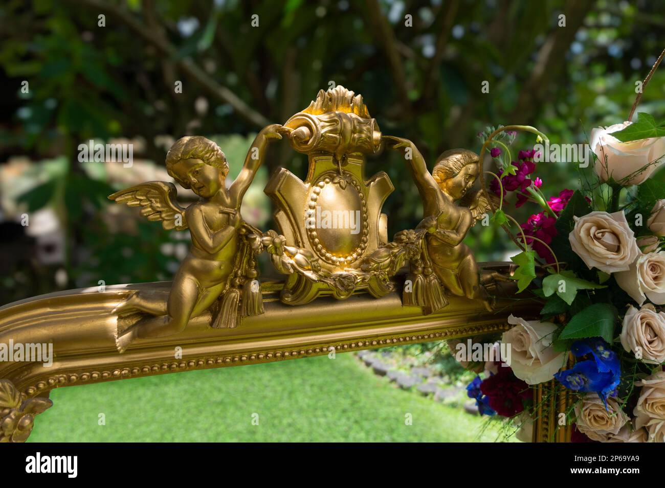 Particolare degli angeli dorati di un antico specchio ornato di rose nel mezzo del giardino che riflette l'erba con uno sfondo fuori fuoco tre Foto Stock