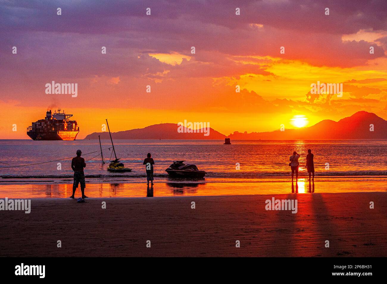 Tramonto con il cielo rossastro sulle spiagge di Santos, São Paulo, Brasile. Nave da carico e persone che contemplano il mare. Foto Stock