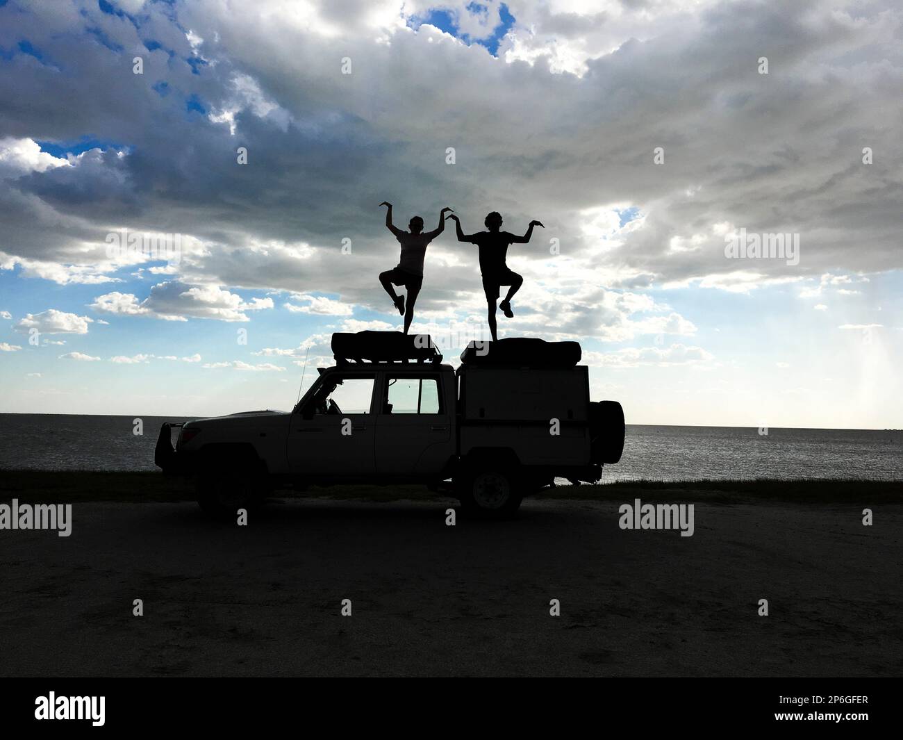 2 giovani adulti in piedi su una jeep come silhouette che eseguono figure umane. Nuvole tempesta nel cielo. Makgadikgadi Salt Pans, Botswana, Africa Foto Stock
