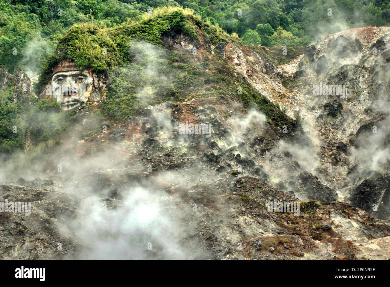 Un campo di fumarole, sullo sfondo di una gigantesca formazione del volto che illustra il carattere del Toar, che, insieme a Lumimuut Sono creduti come gli antenati del popolo di Minahasan secondo la mitologia locale, a Bukit Kasih, una destinazione popolare per il turismo culturale, naturale e religioso situato nel villaggio di Kanonang, Kawangkoan occidentale, Minahasa, Sulawesi settentrionale, Indonesia. Foto Stock