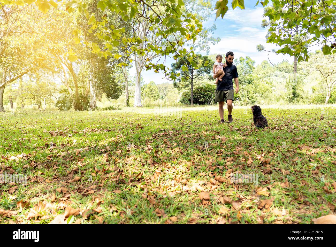 Padre che tiene il suo bambino a camminare con il suo cane nel campo circondato da alberi. Concetto di Father's Day Foto Stock