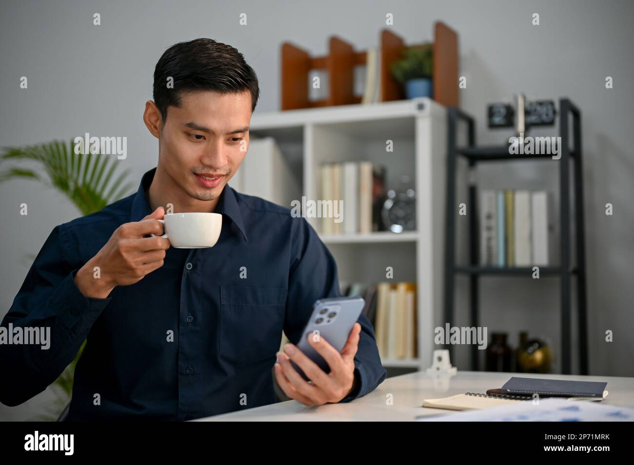 Felice e bello uomo d'affari asiatico millenario utilizzando il suo smartphone per leggere un articolo online sul sito web mentre sorseggiando caffè mattutino in ufficio. Foto Stock