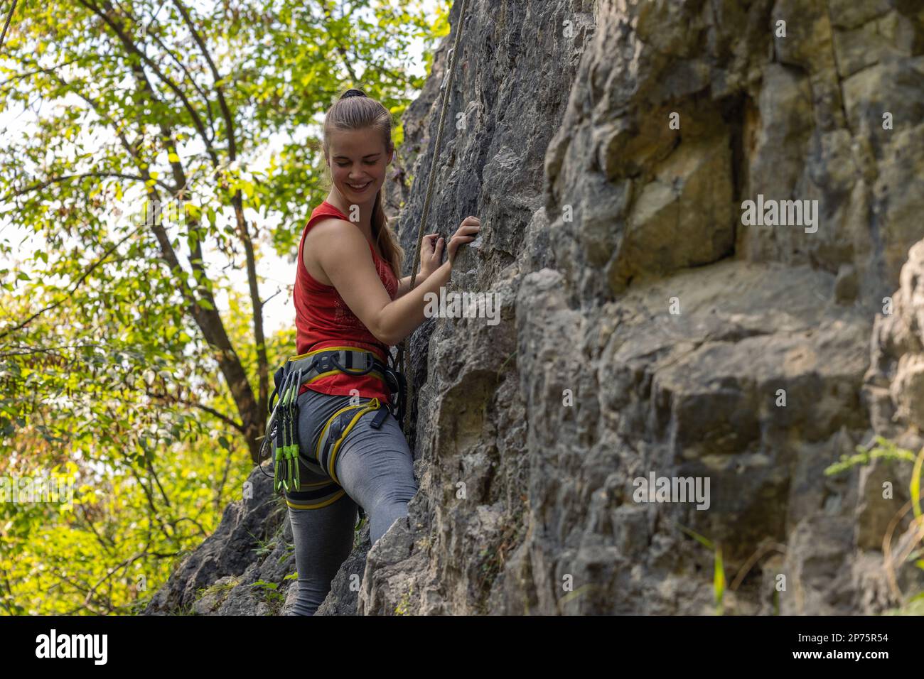 Donna arrampicatrice sorridente e guardando verso il basso mentre si arrampica nella parete rocciosa Foto Stock