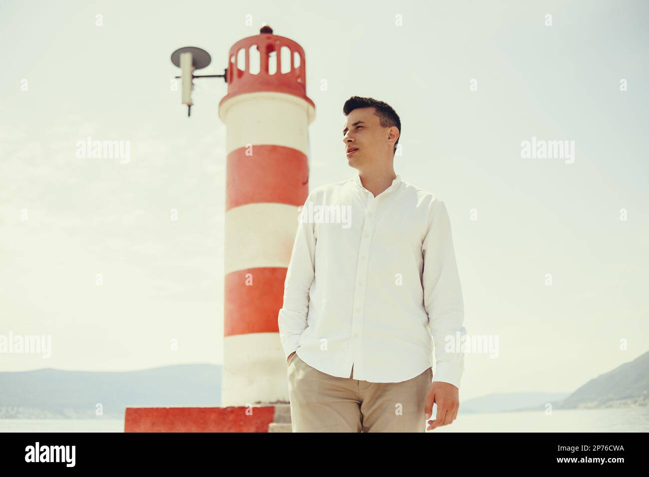 Un uomo in camicia bianca cammina lungo la spiaggia. Il ragazzo sullo sfondo delle montagne e del mare va al faro rosso. Riposo e viaggio Foto Stock