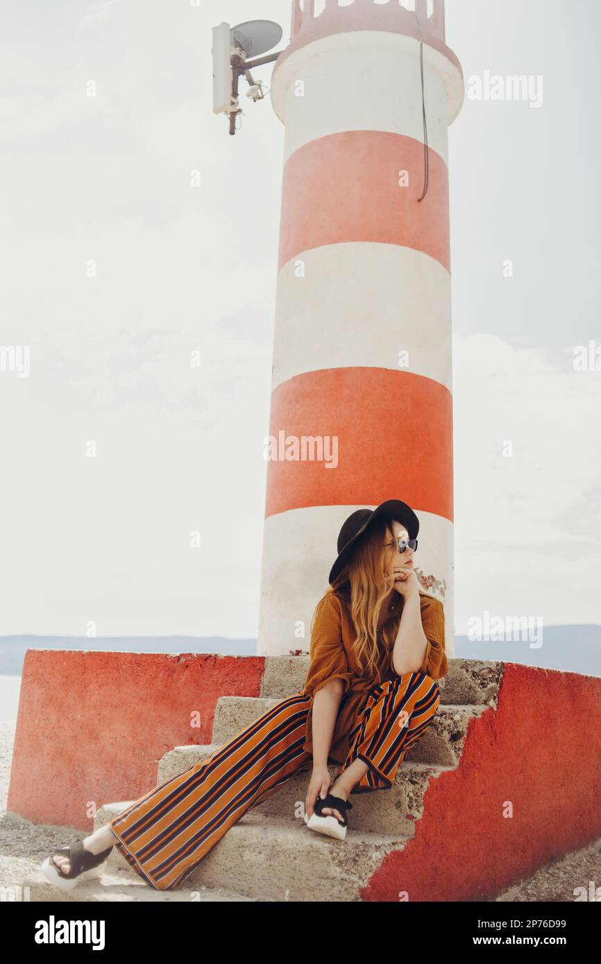 Splendida donna alla moda in posa mentre si siede su un faro. Foto estiva di una ragazza in tuta giallo-arancione, cappello nero e occhiali da sole. Ragazza bionda Foto Stock