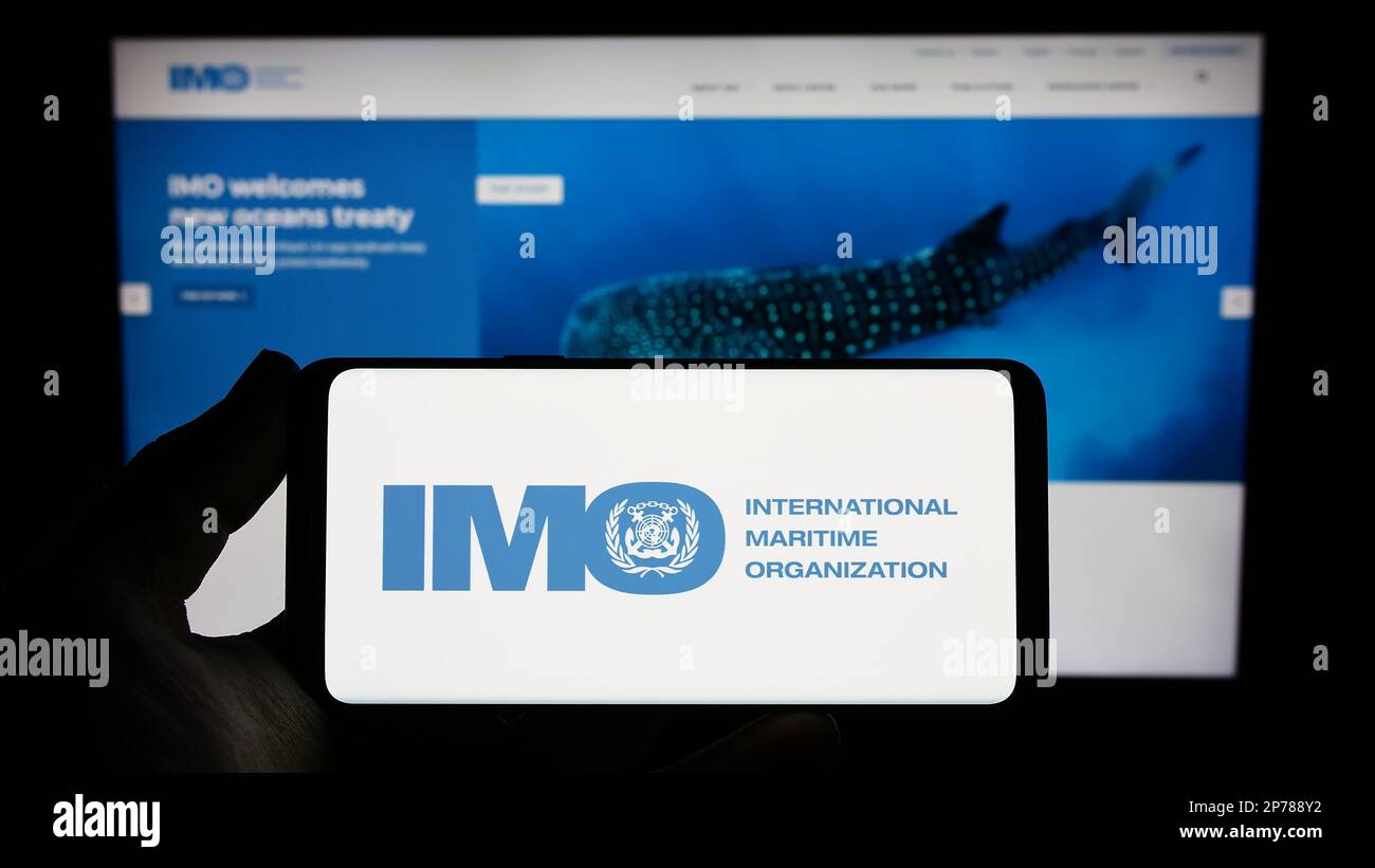 Persona che tiene lo smartphone con il logo dell'Organizzazione marittima internazionale (IMO) sullo schermo davanti al sito Web. Messa a fuoco sul display del telefono. Foto Stock