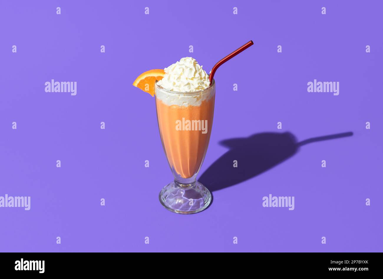 Bicchiere di milkshake arancione sormontato da panna montata minimalista su un tavolo viola. Singolo bicchiere di milkshake in luce brillante isolato su un vivace colo Foto Stock