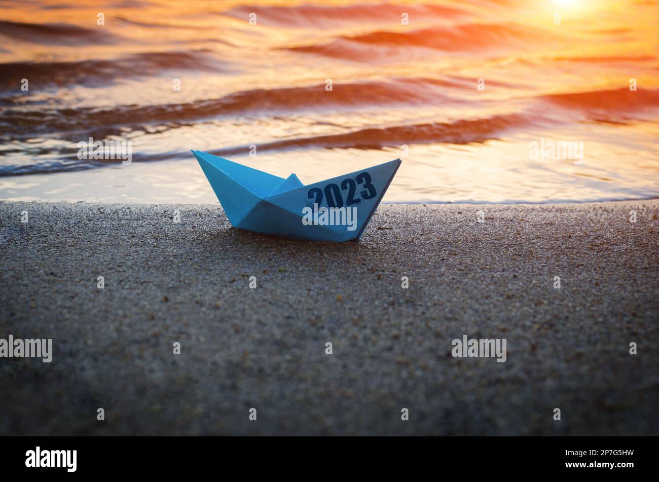 Una barca di carta blu con l'iscrizione a bordo 2023, lavata sulla spiaggia, sullo sfondo dell'oceano e del tramonto. Foto Stock