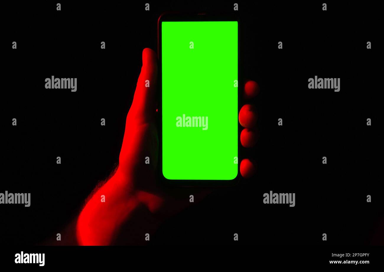 Telefono cellulare in mano da giovane. Utilizzare lo smartphone con schermo verde con illuminazione al neon blu rossa e sfondo scuro. Maschio che mostra nero telefono chroma chiave displ Foto Stock