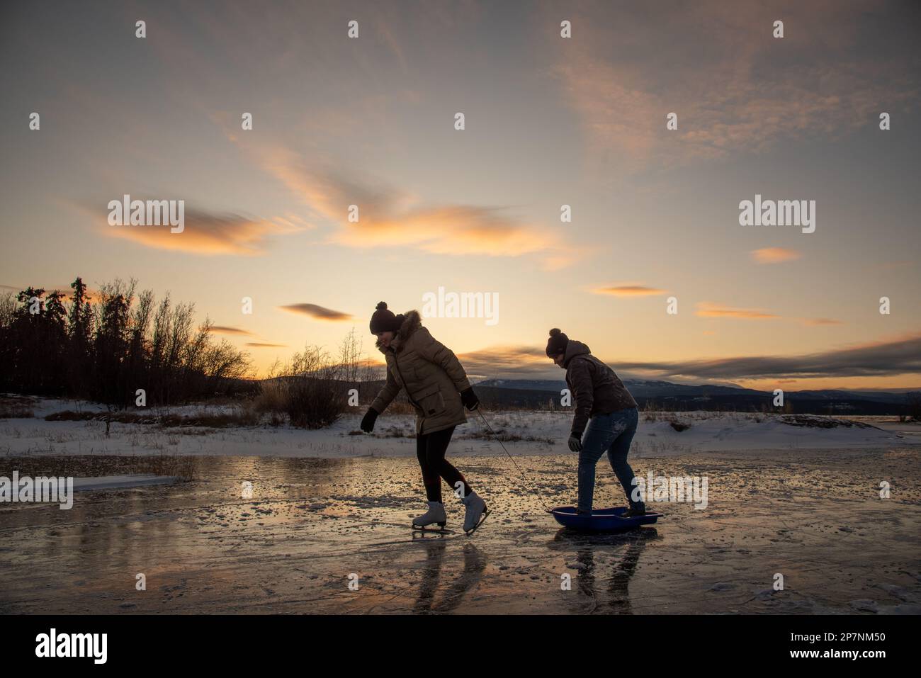 Due donne pattinano sul ghiaccio su un lago selvaggio nel Canada settentrionale all'inizio della stagione invernale con uno splendido sfondo arancione al tramonto. Foto Stock