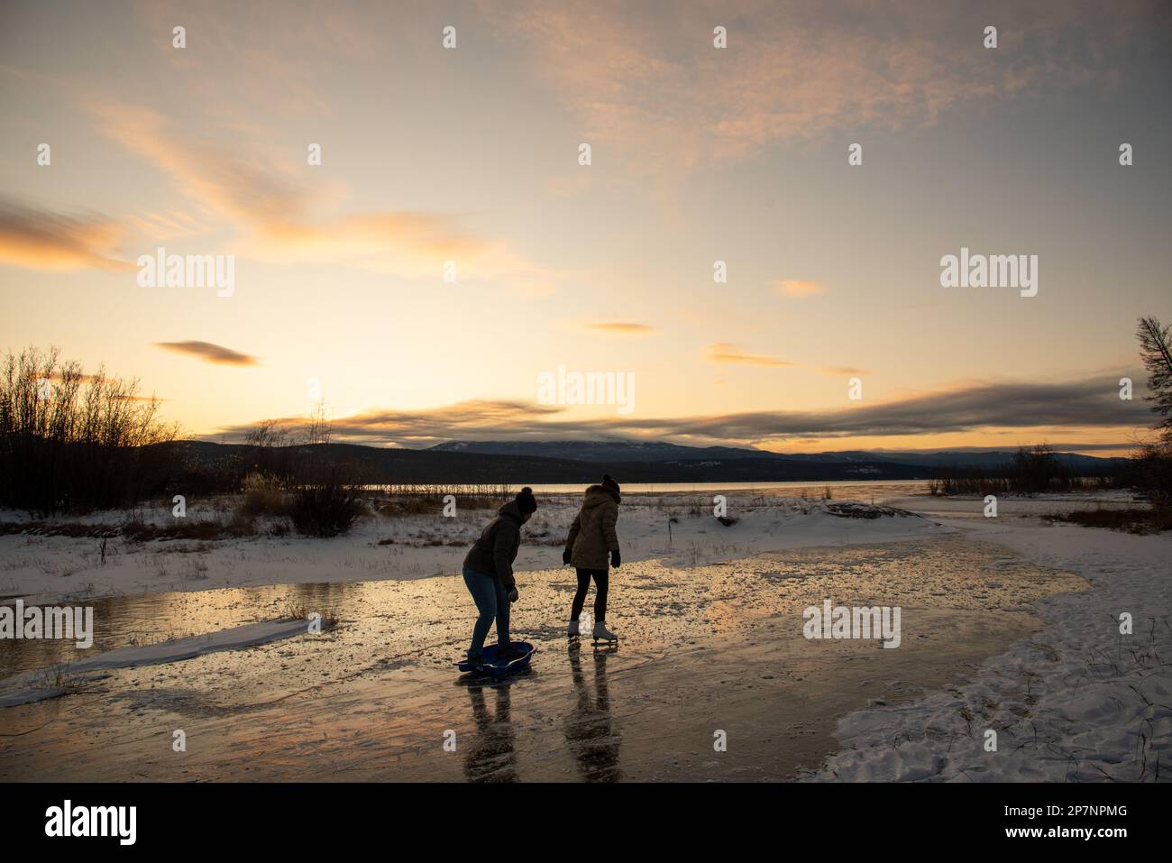 Due donne pattinano sul ghiaccio su un lago selvaggio nel Canada settentrionale all'inizio della stagione invernale con uno splendido sfondo arancione al tramonto. Foto Stock