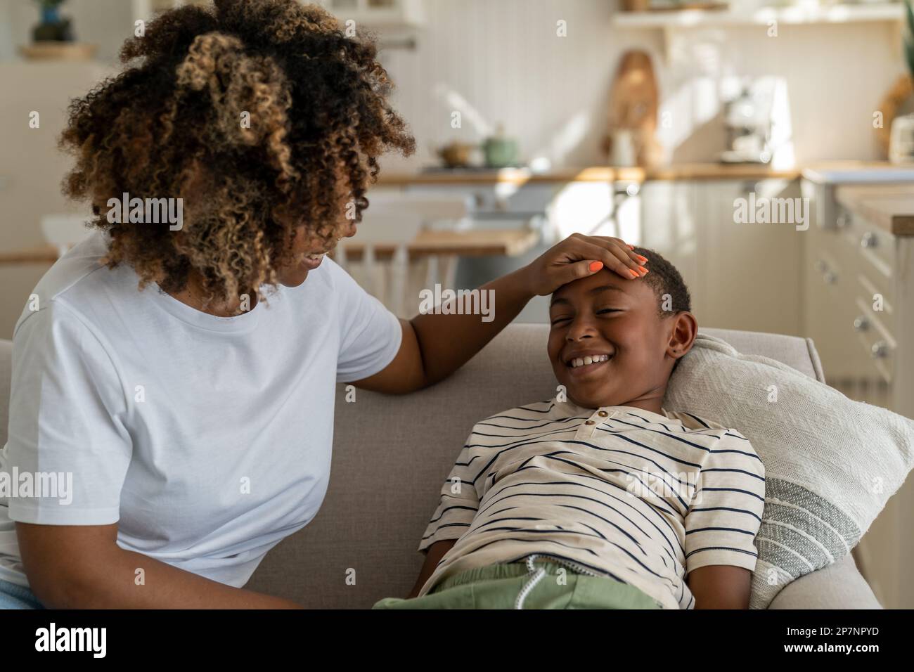 La mamma afro-americana si accosta alla testa e loda sorridendo contentissimo figlio ragazzo seduto sul divano Foto Stock