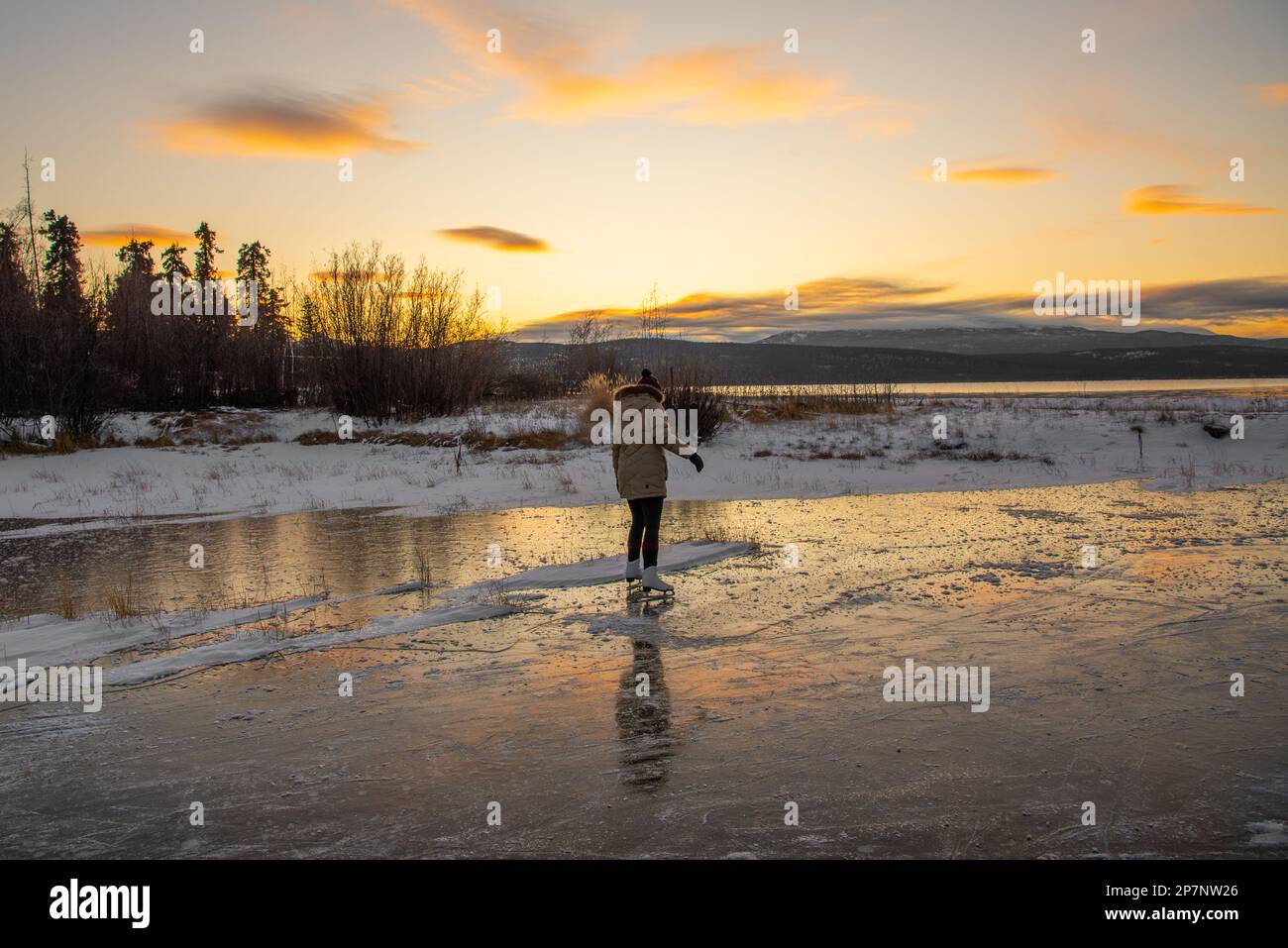 Una donna che pattina su un lago selvaggio nel nord del Canada all'inizio della stagione invernale con uno splendido sfondo arancione tramonto. Foto Stock