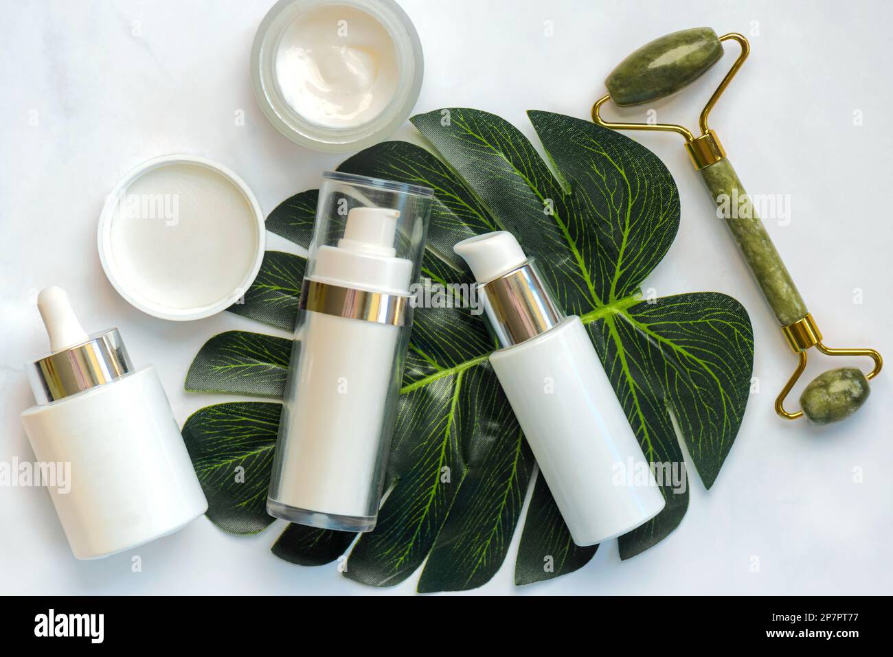 Set di cosmetici e rullo massaggiatore in giada per la cura della pelle con foglia verde su un tavolo in marmo. Concetto di prodotti per la cura della pelle Foto Stock