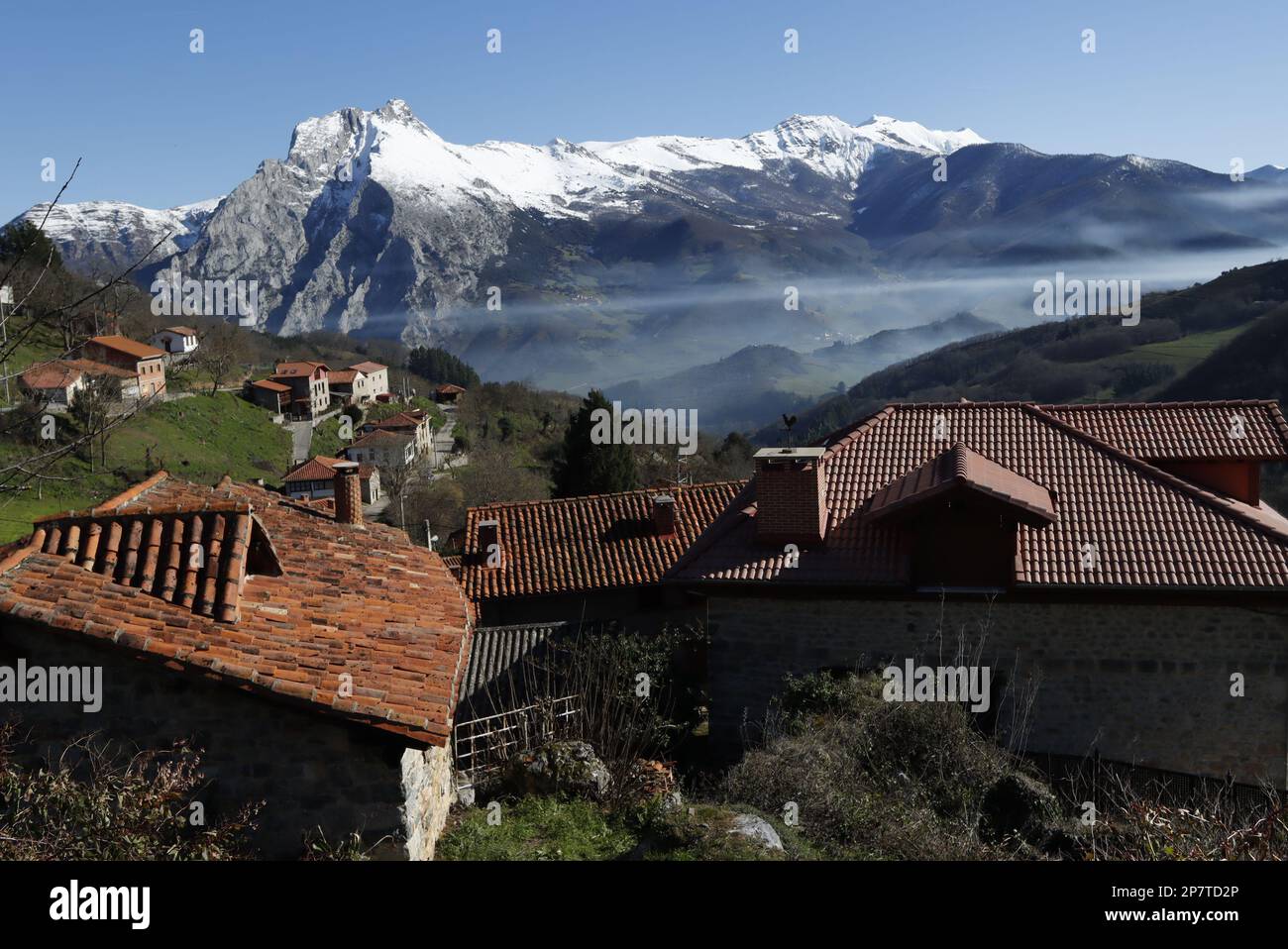 Tejados de teja roja y casa del pueblo de Colio en el valle con niebla de Liébana, con sus prados verdes y la montaña nevada que los cirunda.. Foto Stock