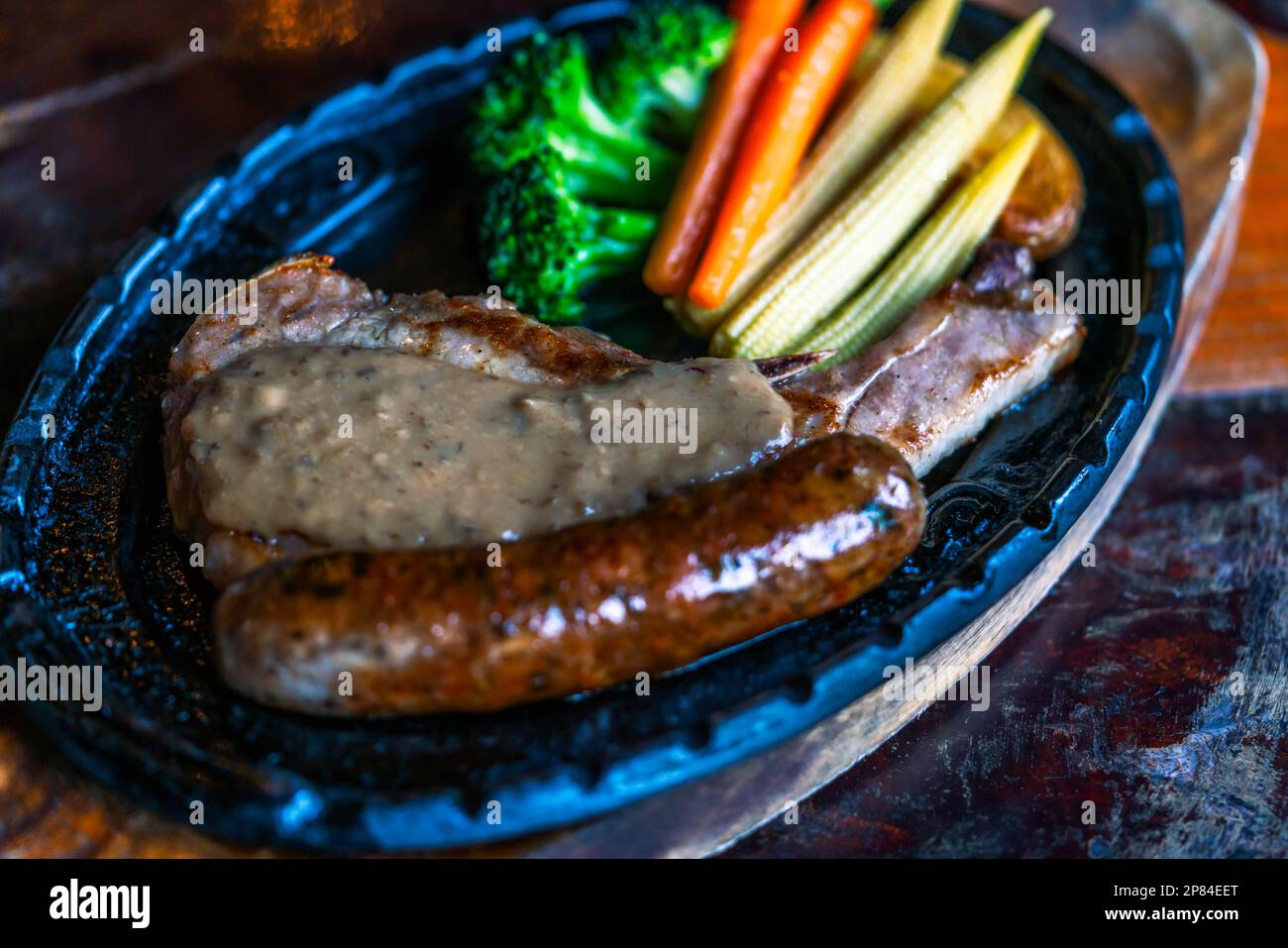 Primo piano trita di maiale bistecca di pepe, salsiccia, patate e verdure su piastra di ferro caldo, vecchio tavolo di legno, luce naturale. Foto Stock