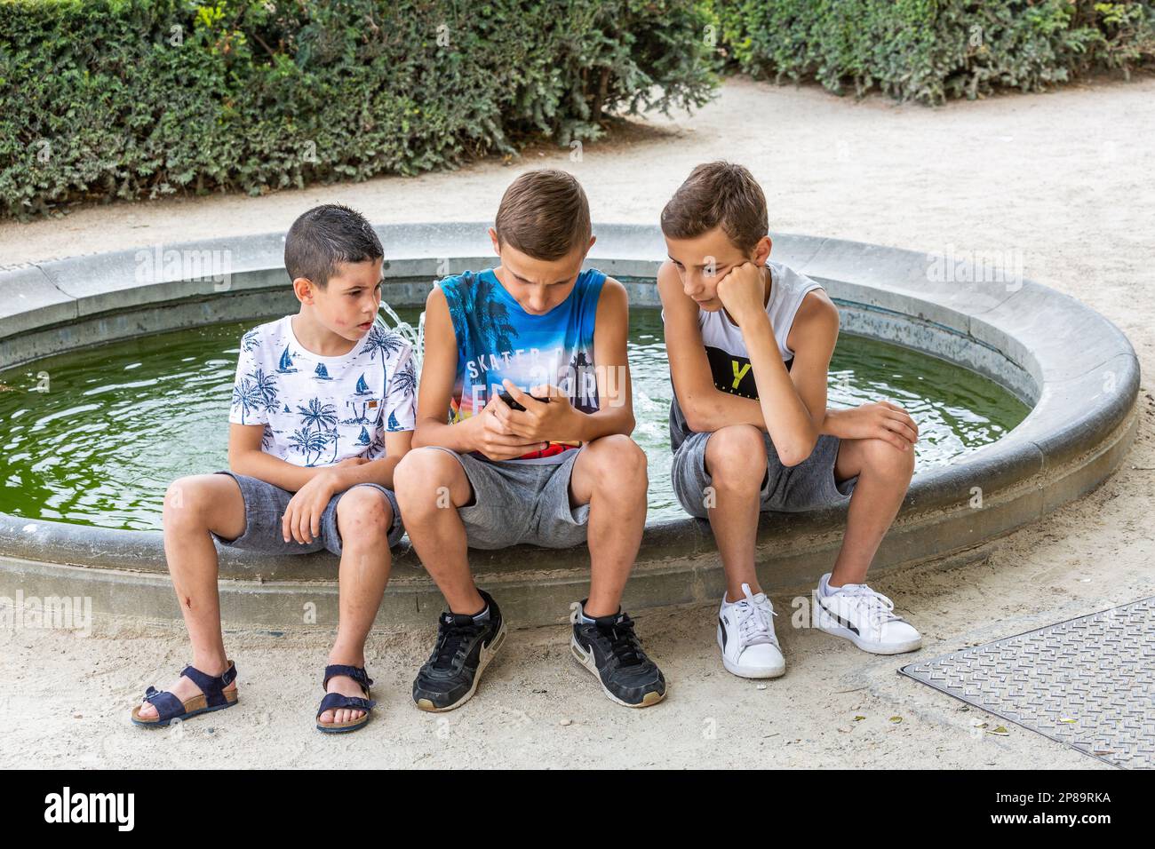 Tre ragazzi, seduti sul bordo del bacino di una fontana, guardando lo smartphone che uno di loro sta usando. Bruxelles. Foto Stock