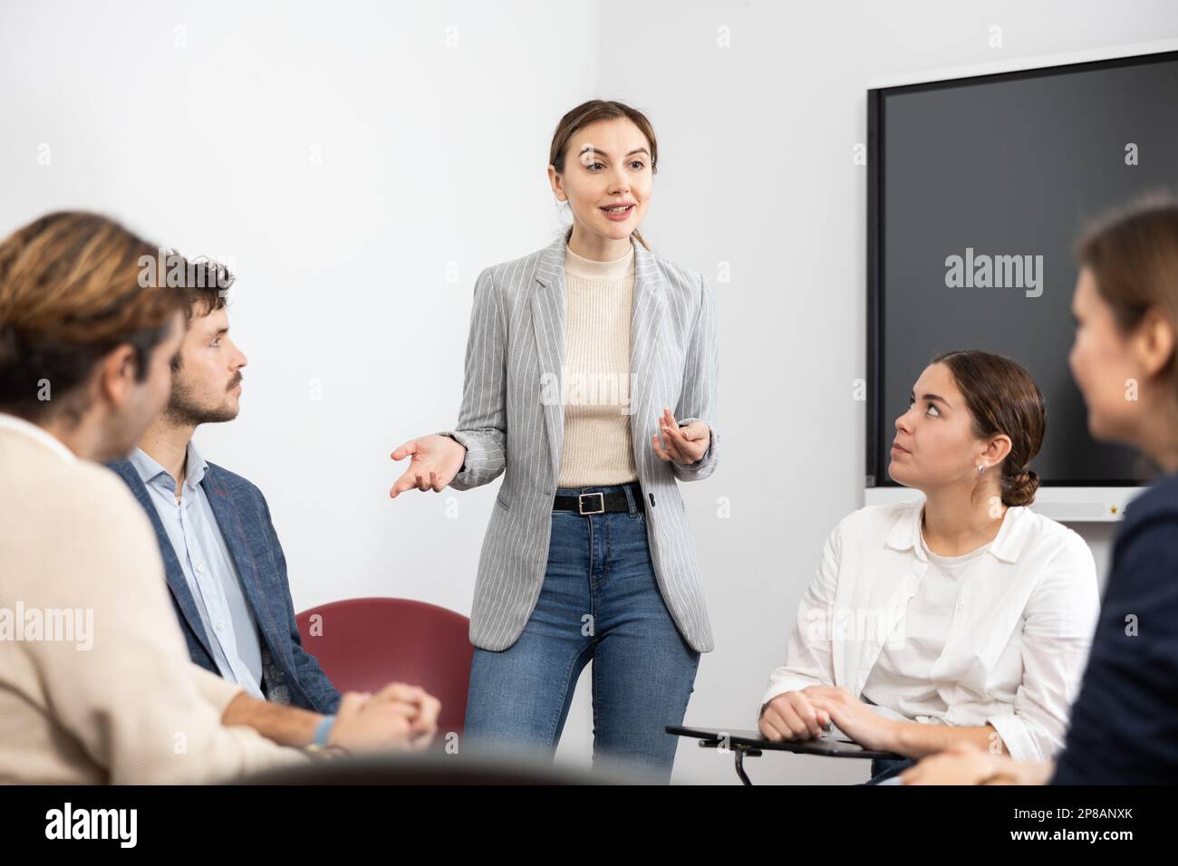 Allenatore professionale donna in piedi vicino a tavola interattiva e condurre formazione diverse persone riunite in cerchio in auditorium Foto Stock
