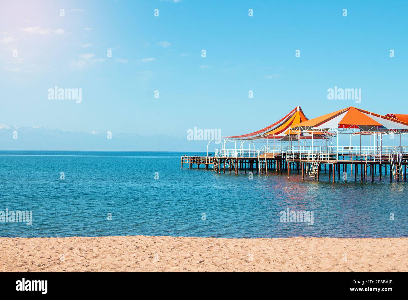 Molo in legno con tendone rosso-arancio su acqua di mare, lago, oceano con spiaggia senza persone. Viaggi, vacanze, sfondo estivo. Spazio di copia Foto Stock