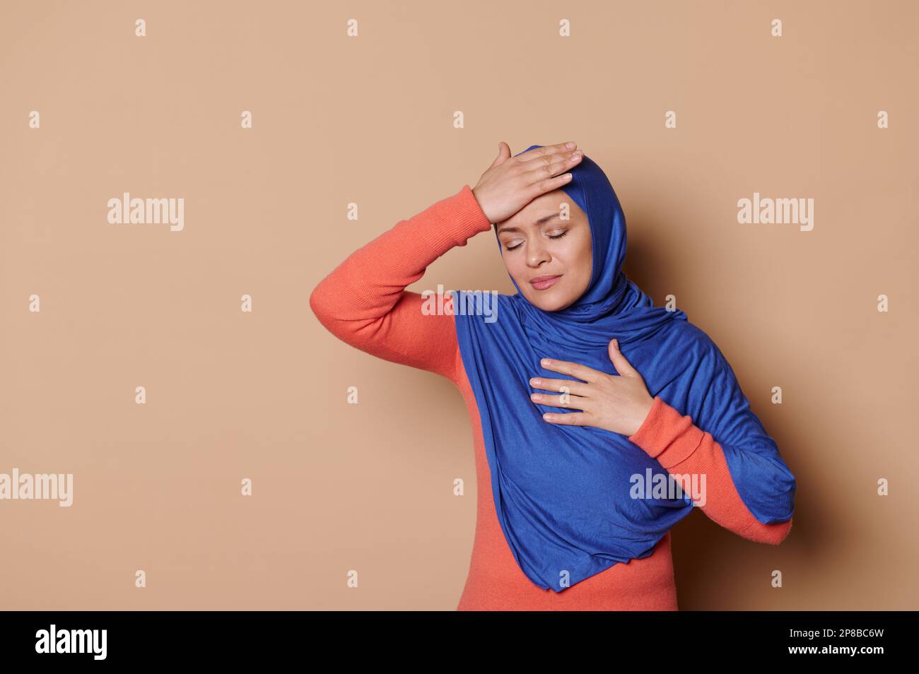Donna musulmana in hijab blu, tenendo la mano sulla testa, che soffre di mal di testa, terribile emicrania, sfondo beige isolato Foto Stock